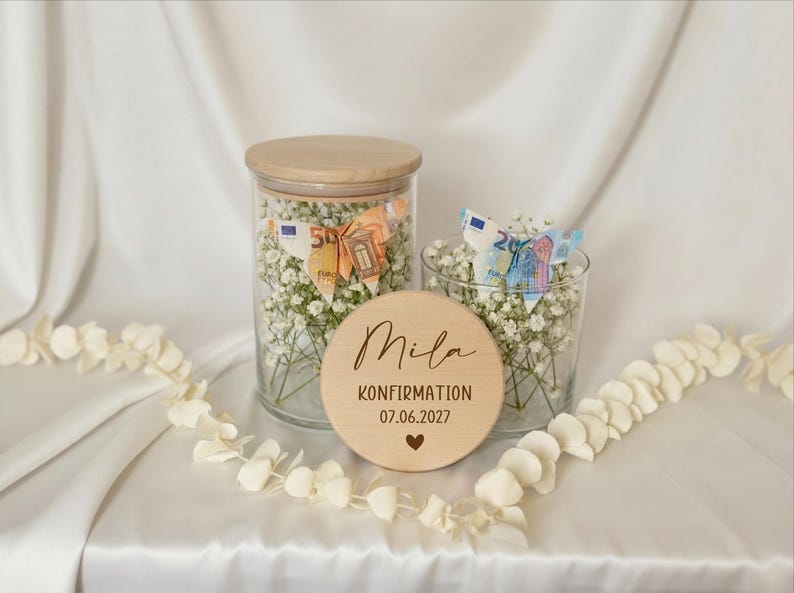 May include: Two glass jars filled with white flowers and money, one with a wooden lid. A wooden disc with the name "Mila", the word "KONFIRMATION", the date "07.06.2027", and a heart symbol is in front of the jars. A white floral garland is in the foreground.