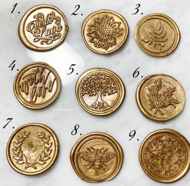 Op de afbeelding: Negen gouden waszegels met verschillende ontwerpen. De ontwerpen omvatten een ​​zonnebloem, een boom, een bij, bladeren en de woorden "with love".