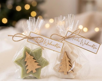 Puede incluir: Dos jabones en forma de estrella empaquetados en bolsas de plástico transparente, uno de color verde salvia y otro blanco roto. Cada jabón tiene un recorte de árbol de Navidad de madera y una etiqueta de papel con Frohe Weihnachten.
