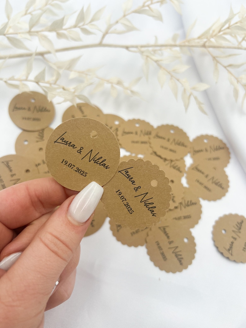 May include: Brown paper circle tags with scalloped edges. The tags have a hole punched in the top and are decorated with black text that reads "Laura & Niklas" and the date "19.07.2025".