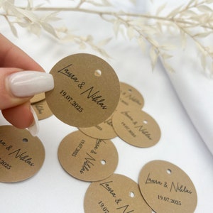 May include: Brown circular paper tags with the text "Laura & Niklas" and the date "19.07.2025" printed in black ink. The tags have a hole punched in the top.