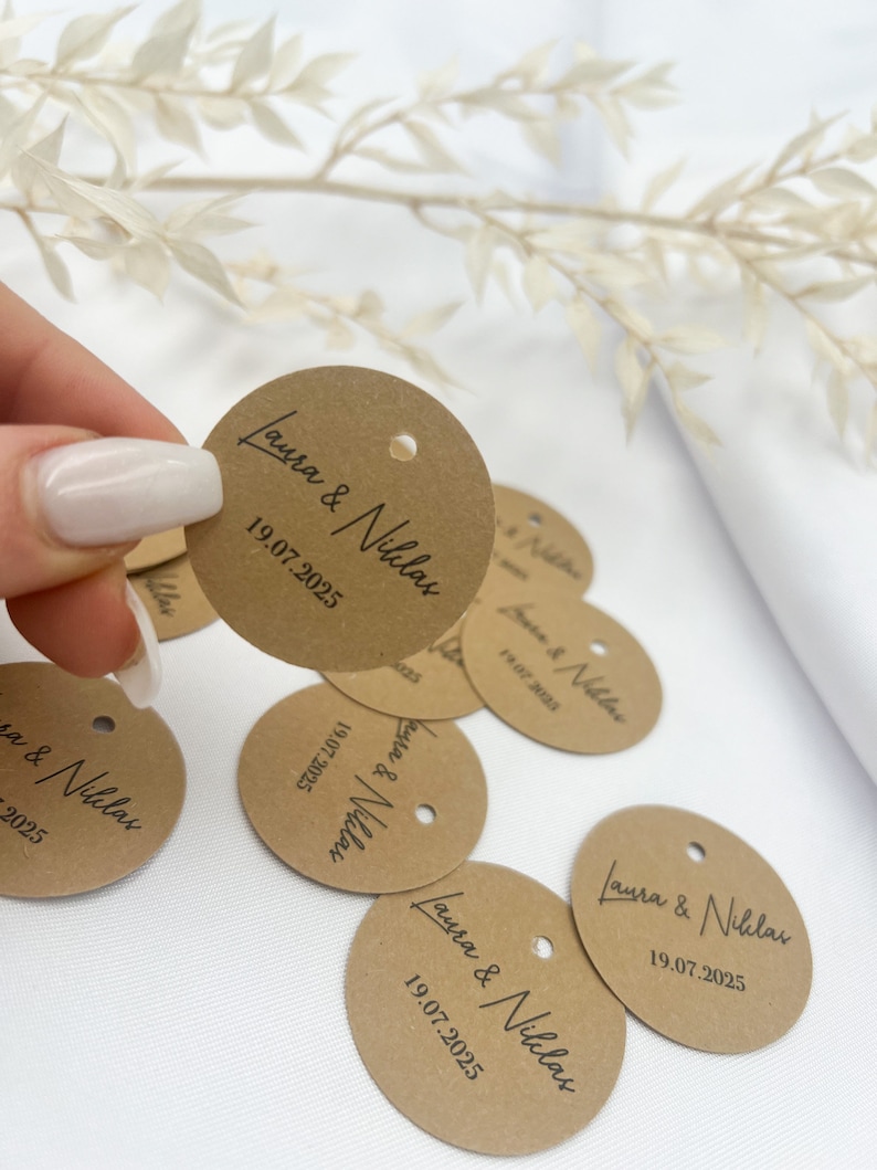May include: Brown circular paper tags with the text "Laura & Niklas" and the date "19.07.2025" printed in black ink. The tags have a hole punched in the top.