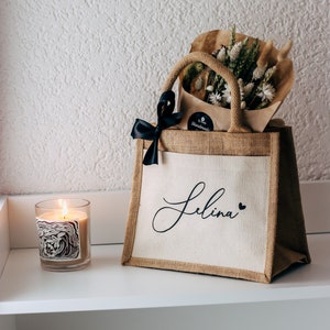 May include: A brown burlap tote bag with a white canvas insert. The bag has a black ribbon bow and the name "Selina" written in black cursive on the front. The bag is filled with a bouquet of dried flowers.