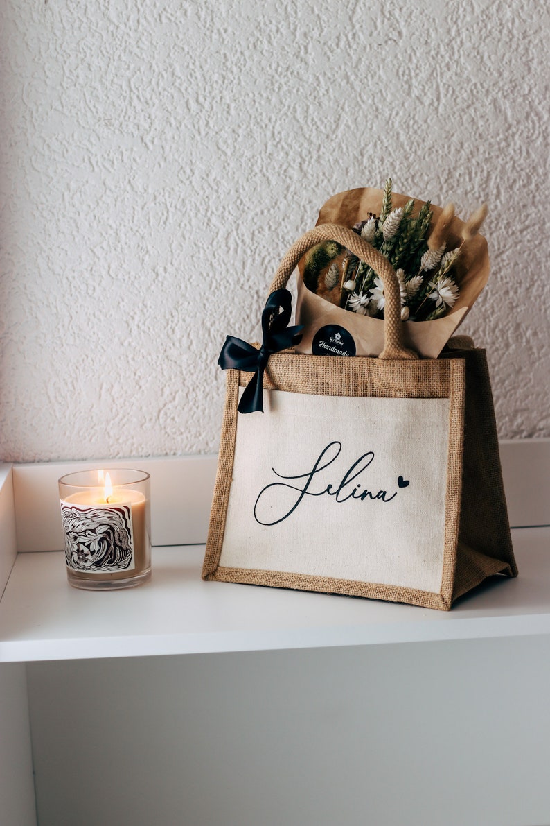 May include: A brown burlap tote bag with a white canvas insert. The bag has a black ribbon bow and the name "Selina" written in black cursive on the front. The bag is filled with a bouquet of dried flowers.