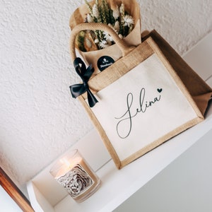May include: A brown burlap tote bag with a white canvas insert. The bag has a black ribbon bow and a black sticker that says "Handmade". The canvas insert has the name "Selina" written in black cursive with a small black heart. The bag is filled with dried flowers and is sitting on a white shelf with a candle.