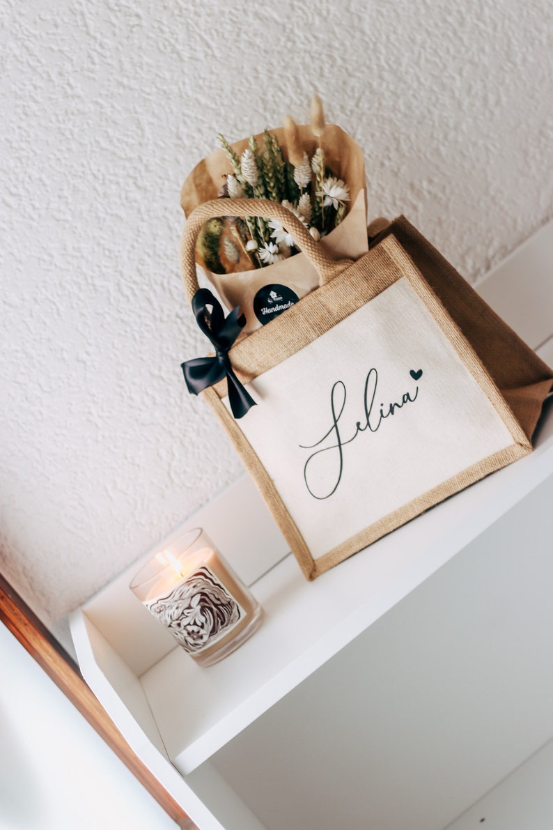 May include: A brown burlap tote bag with a white canvas insert. The bag has a black ribbon bow and a black sticker that says "Handmade". The canvas insert has the name "Selina" written in black cursive with a small black heart. The bag is filled with dried flowers and is sitting on a white shelf with a candle.