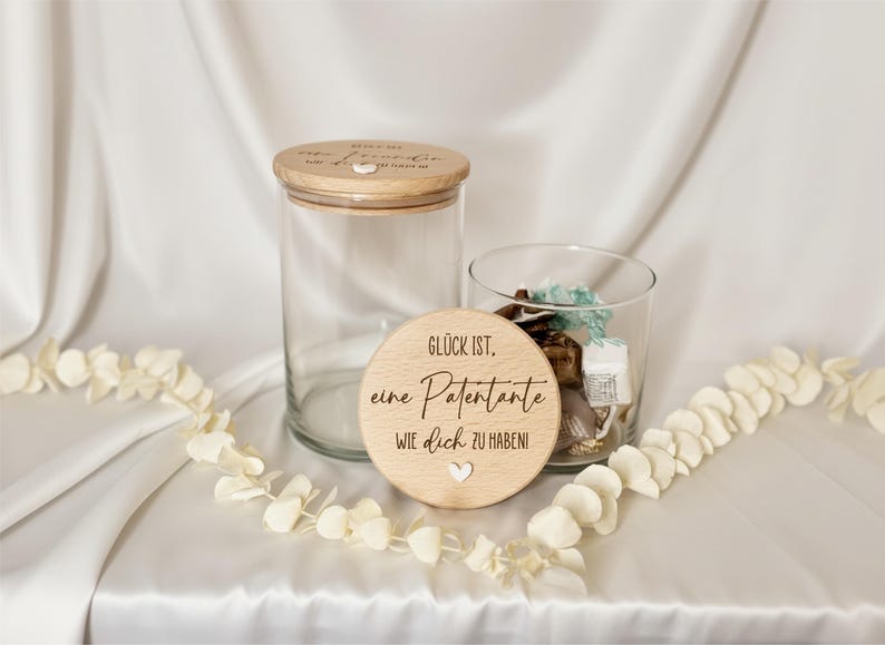 K&ouml;nnte beinhalten: Zwei klare Glasbeh&auml;lter mit Holzdeckeln und ein Holzdeckel mit dem Text "Gl&uuml;ck ist, eine Patentante wie dich zu haben!" Eine wei&szlig;e Blumengirlande ist &uuml;ber die wei&szlig;e Oberfl&auml;che drapiert.