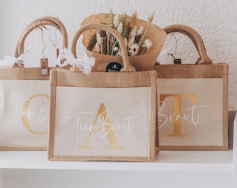 May include: Three brown burlap tote bags with white lettering and gold foil accents. The bags are decorated with the words "Team Braut" and "Braut".