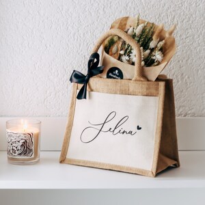 May include: A personalized jute tote bag with a black ribbon and the name "Selina" printed on the front. The bag is filled with dried flowers and is sitting on a white shelf.