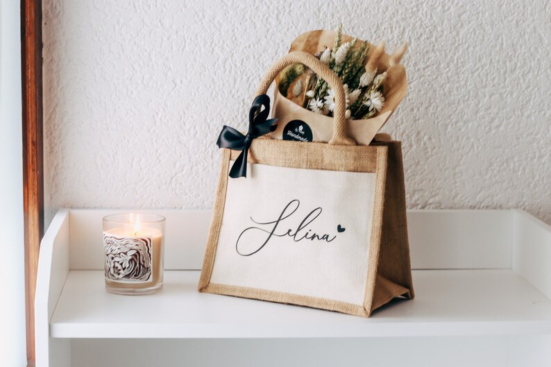 May include: A personalized jute tote bag with a black ribbon and the name "Selina" printed on the front. The bag is filled with dried flowers and is sitting on a white shelf.