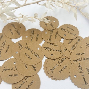 May include: A collection of brown paper circle tags with scalloped edges and a hole punched in the top. The tags are decorated with black calligraphy that reads "Laura & Niklas" and the date "19.07.2025".