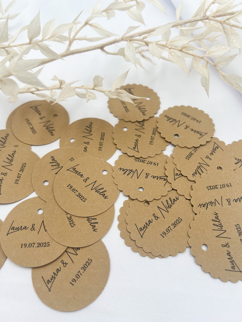 May include: A collection of brown paper circle tags with scalloped edges and a hole punched in the top. The tags are decorated with black calligraphy that reads "Laura & Niklas" and the date "19.07.2025".