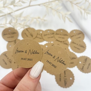 May include: Brown paper circle tags with scalloped edges and a hole for string. The tags have the text "Laura & Niklas" and the date "19.07.2025" printed in black.