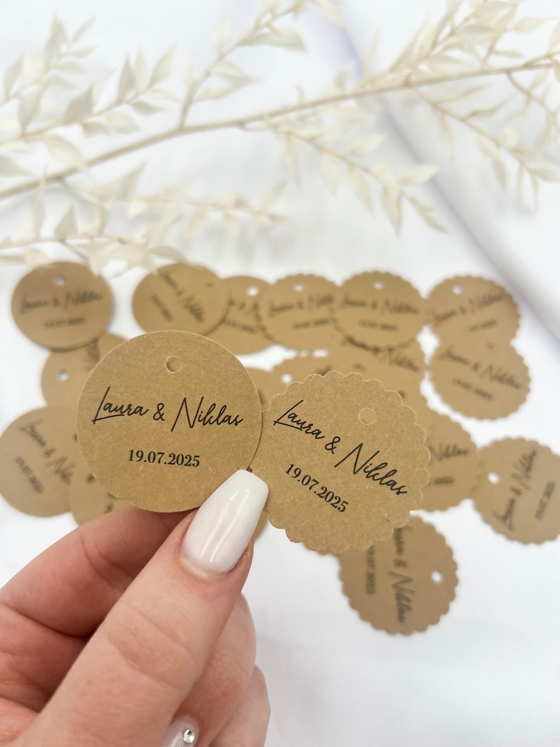 May include: Brown paper circle tags with scalloped edges and a hole for string. The tags have the text "Laura & Niklas" and the date "19.07.2025" printed in black.