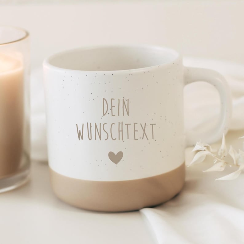 May include: A white ceramic mug with a speckled finish and a tan base. The mug features the text "DEIN WUNSCHTEXT" in light brown, along with a small heart symbol. A candle in a glass holder is visible in the background.