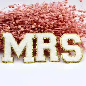 Op de afbeelding: Witte viltletters die "MRS" spellen met een gouden glitterrand. De letters zijn op een witte achtergrond met een boeket roze gedroogde bloemen.