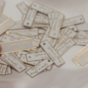 May include: A close-up of a white rectangular tag with gold trim and the text "Sara & Jamie 10.12.2022" in gold lettering. The tag is being held by a finger. There are many similar tags scattered on a white surface.