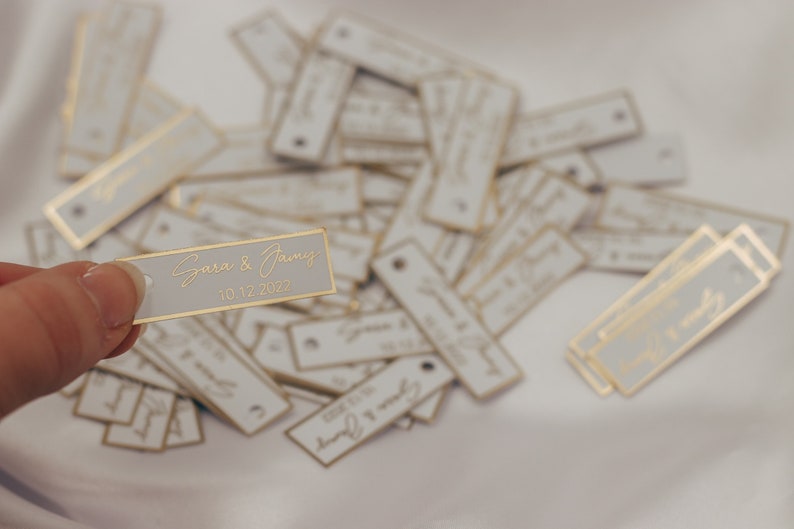 May include: A close-up of a white rectangular tag with gold trim and the text "Sara & Jamie 10.12.2022" in gold lettering. The tag is being held by a finger. There are many similar tags scattered on a white surface.