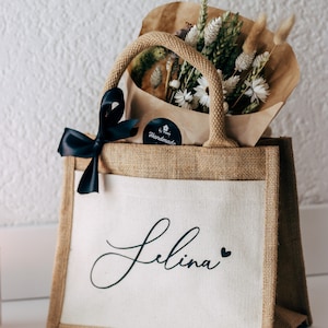 May include: A brown burlap tote bag with a white canvas front. The bag has black handles and a black ribbon tied around the handles. The bag has a black and white sticker that says "Handmade" and the name "Selina" is printed in black cursive on the front of the bag. The bag is filled with a bouquet of dried flowers.