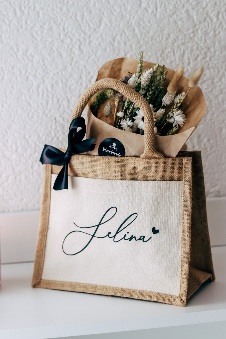 May include: A brown burlap tote bag with a white canvas front. The bag has black handles and a black ribbon tied around the handles. The bag has a black and white sticker that says "Handmade" and the name "Selina" is printed in black cursive on the front of the bag. The bag is filled with a bouquet of dried flowers.