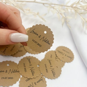 May include: Brown paper circle tags with scalloped edges and a hole punched at the top. The tags have the text "Laura & Niklas" and the date "19.07.2025" printed in black.