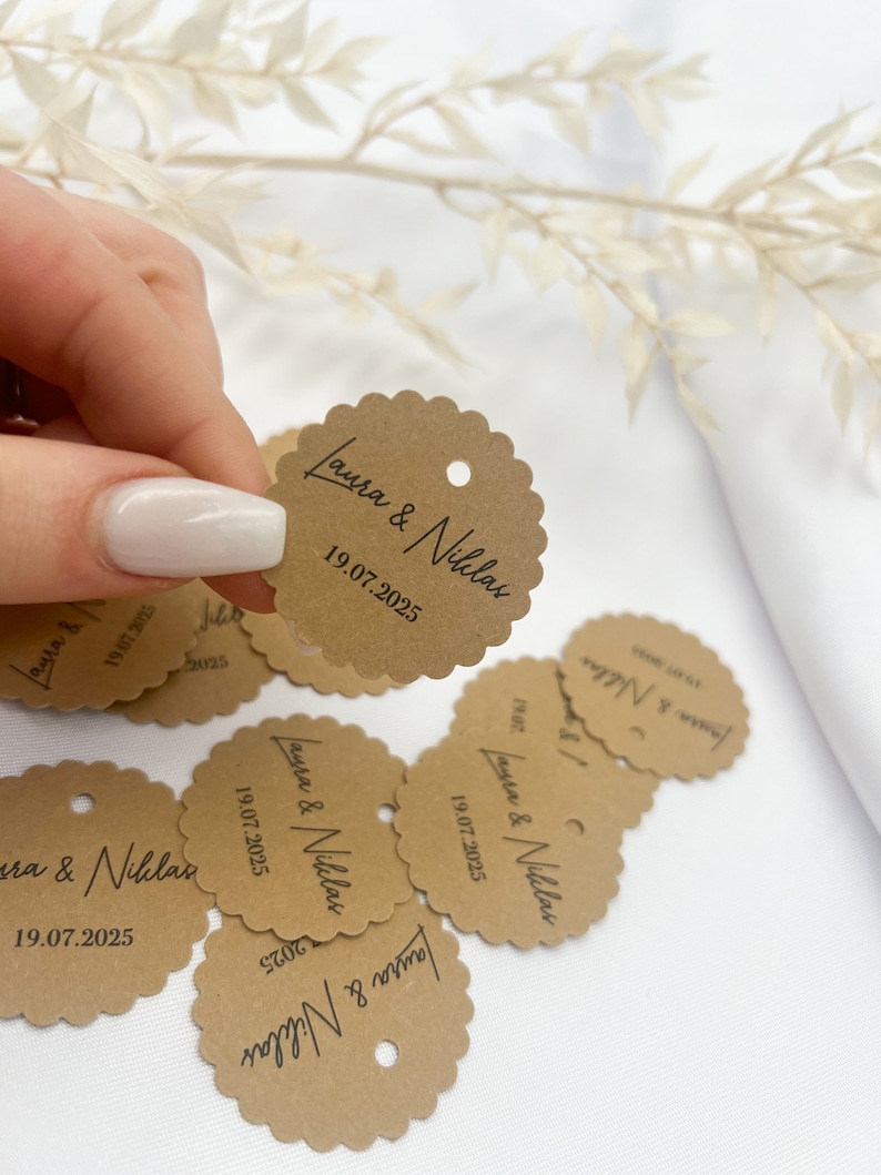 May include: Brown paper circle tags with scalloped edges and a hole punched at the top. The tags have the text "Laura & Niklas" and the date "19.07.2025" printed in black.