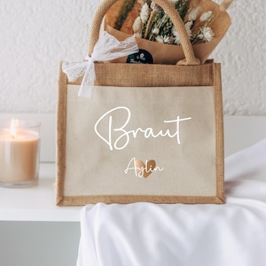 Könnte beinhalten: Eine Jute-Tragetasche mit einem weißen Canvas-Einsatz. Die Tasche hat eine weiße Schleife aus Spitze und eine weiße Aufschrift mit der Aufschrift "Braut Aylin" mit einem goldenen Herzen.