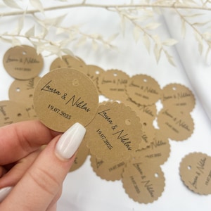 May include: Brown paper circle tags with scalloped edges and a hole punched in the center. The tags are printed with the text "Laura & Nicolas" and the date "19.07.2025".