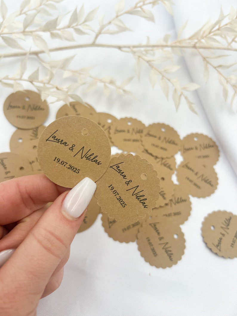May include: Brown paper circle tags with scalloped edges and a hole punched in the center. The tags are printed with the text "Laura & Nicolas" and the date "19.07.2025".