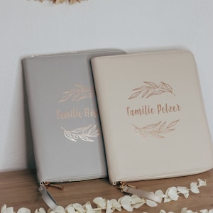 May include: Two gray and beige zippered pouches with gold foil lettering. The pouches have a floral design and the text "Familie Floß" and "Familie Pelzer".