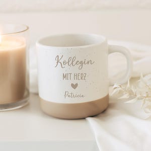 May include: A white ceramic mug with a speckled design and a tan base. The mug features the text "Kollegin MIT HERZ" with a heart symbol and the name "Patricia". A lit candle is visible in the background.