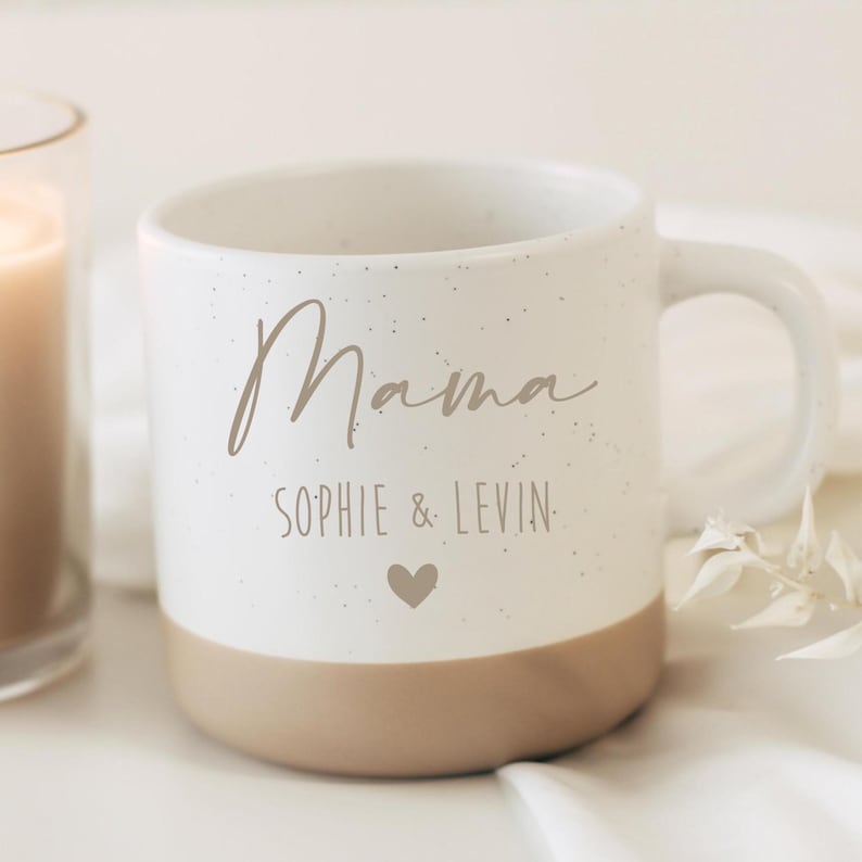 Pode incluir: Uma caneca de cer&acirc;mica com uma se&ccedil;&atilde;o superior branca salpicada e uma base bege s&oacute;lida. A frente apresenta a palavra "Mama" em letra cursiva, seguida pelo texto "SOPHIE & LEVIN" em fonte sem serifa e um pequeno &iacute;cone de cora&ccedil;&atilde;o abaixo.