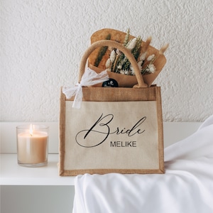 Könnte beinhalten: Eine braune Jute-Tragetasche mit weißer Spitzenverzierung und dem Text "Bride Melike" in schwarzer Schrift. Die Tasche ist mit getrockneten Blumen und braunem Geschenkpapier gefüllt.