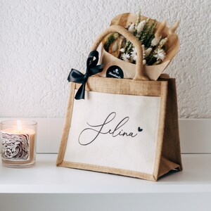 May include: A brown burlap tote bag with a white fabric insert. The bag has a black ribbon bow and the name "Selina" written in black cursive with a small black heart next to it. The bag is filled with dried flowers and a small brown paper bag.