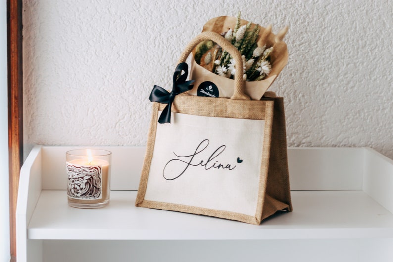 May include: A brown burlap tote bag with a white fabric insert. The bag has a black ribbon bow and the name "Selina" written in black cursive with a small black heart next to it. The bag is filled with dried flowers and a small brown paper bag.