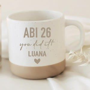 Könnte beinhalten: Ein Keramikbecher mit gesprenkeltem weißem Oberteil und einem einfarbigen beigen Boden. Auf der Vorderseite steht der Text "ABI 26", "you did it!" und "LUANA" in brauner Schrift, ergänzt durch ein kleines braunes Herz unter dem Namen.