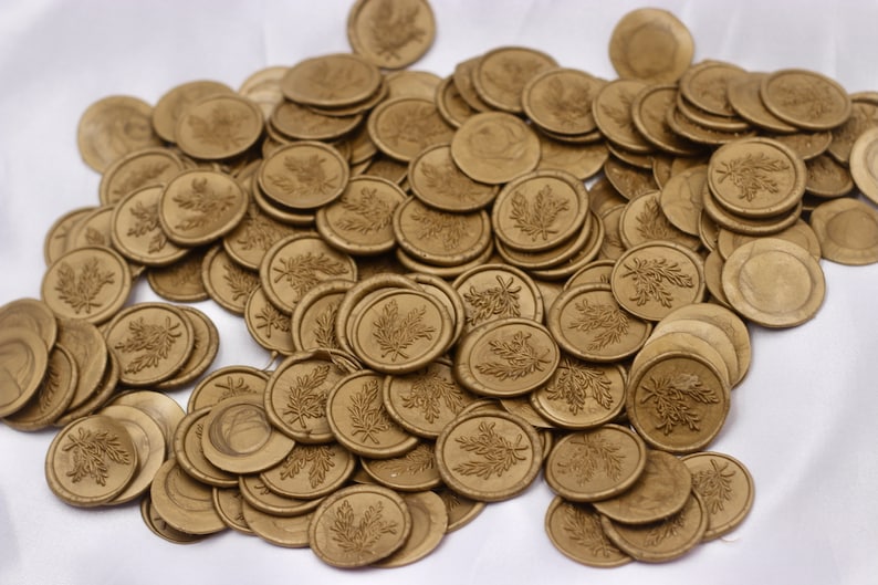 Op de afbeelding: Een grote stapel gouden waszegels met een bladerenontwerp. De zegels zijn verspreid over een wit oppervlak.