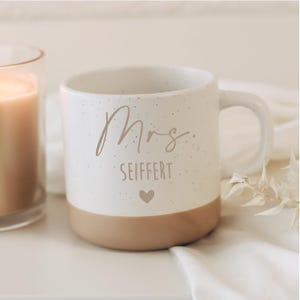 Könnte beinhalten: Eine weiße Keramiktasse mit gesprenkelter Oberfläche und beigem Boden. Die Tasse trägt den Schriftzug "Mrs." in Schreibschrift, den Namen "SEIFFERT" in einer serifenlosen Schriftart und ein kleines Herzsymbol.