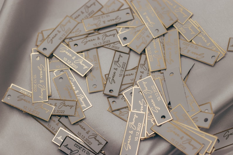 May include: A pile of gray rectangular tags with gold trim and the text "Sara & Jamy 10.12.2022" printed on them.
