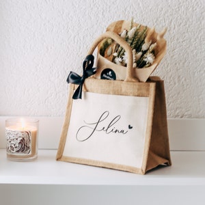 May include: A personalized jute tote bag with a black ribbon and a bouquet of dried flowers. The bag is decorated with the name "Selina" in cursive script and a small heart.