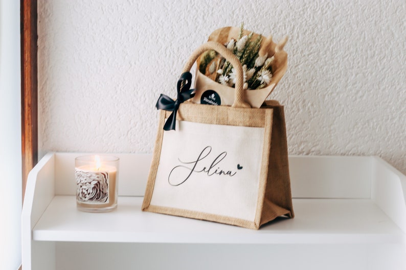May include: A personalized jute tote bag with a black ribbon and a bouquet of dried flowers. The bag is decorated with the name "Selina" in cursive script and a small heart.