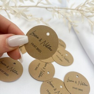 May include: A collection of brown paper circle tags with the text "Laura & Niklas" and the date "19.07.2025" printed in black.