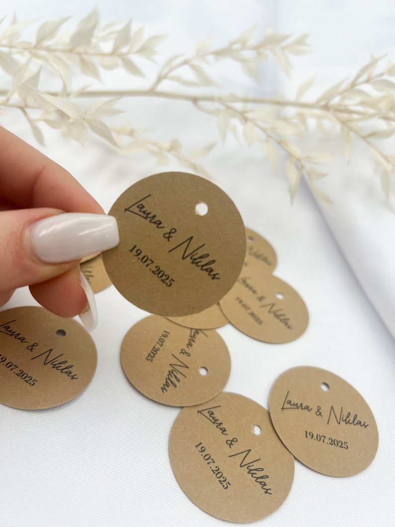 May include: A collection of brown paper circle tags with the text "Laura & Niklas" and the date "19.07.2025" printed in black.
