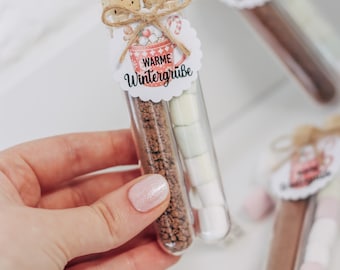 Puede incluir: Tubos transparentes llenos de mezcla de cacao y malvaviscos, rematados con corchos y etiquetas decorativas. Las etiquetas dicen "WARME Wintergrüße" y presentan un gráfico de taza. Una mano sostiene un tubo, con otros en el fondo.