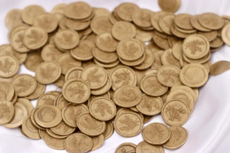 Op de afbeelding: Een stapel gouden waszegels met een bladerenontwerp. De zegels zijn verspreid over een wit oppervlak.