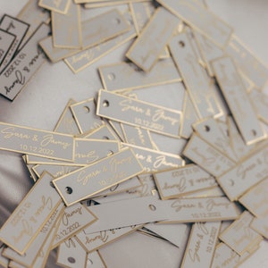 May include: A pile of white rectangular tags with gold trim and the text "Sara & Jamy 10.12.2022" printed on them.