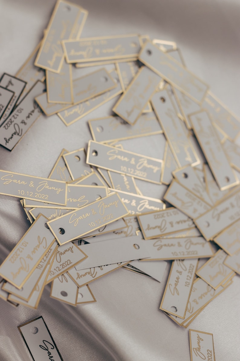 May include: A pile of white rectangular tags with gold trim and the text "Sara & Jamy 10.12.2022" printed on them.