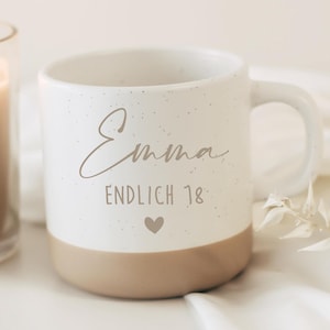 Pu&ograve; includere: Una tazza in ceramica bianca con finitura maculata e base color marrone chiaro. Sul davanti &egrave; riportato il nome Emma in corsivo, seguito dalla scritta ENDLICH 18 e da una piccola icona a forma di cuore di colore marrone. La tazza ha un manico.