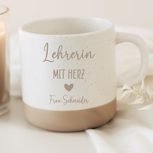 Könnte beinhalten: Eine weiße Keramiktasse mit gesprenkelter Oberfläche und beigem Boden. Auf der Vorderseite steht der Text "Lehrerin MIT HERZ" über einem kleinen Herzsymbol und dem Namen "Frau Schneider" in Schreibschrift. Die Tasse hat einen geschwungenen Henkel.