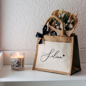 May include: A brown burlap tote bag with a white canvas insert. The bag has a black ribbon bow and the name "Selina" written in black script on the front. The bag is filled with a bouquet of dried flowers and a small brown paper bag with a "Handmade" label.